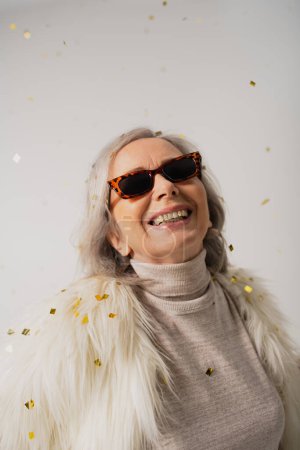 Téléchargez les photos : Femme âgée souriante en fausse fourrure blanche et lunettes de soleil tendance près de confettis tombants sur fond gris - en image libre de droit