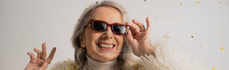 cheerful senior woman adjusting trendy sunglasses near falling confetti on grey background, banner 