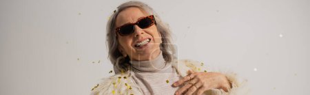 smiling senior woman in white faux fur jacket and trendy sunglasses near falling confetti on grey background, banner 