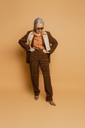 Téléchargez les photos : Full length of stylish senior woman in sunglasses and suit posing with hands on hips on beige - en image libre de droit
