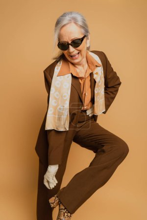 Photo for Cheerful senior woman in sunglasses and suit posing with hand on hip on beige - Royalty Free Image