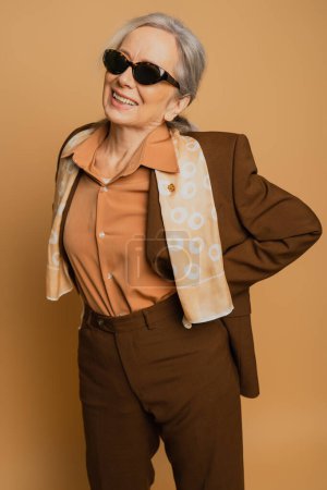 Photo for Positive and senior woman in sunglasses and suit posing with hand on hip on beige - Royalty Free Image
