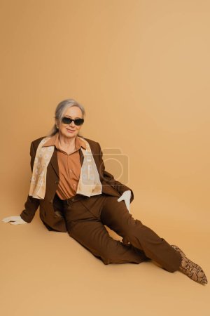 full length of cheerful senior woman in sunglasses and suit sitting on beige background 