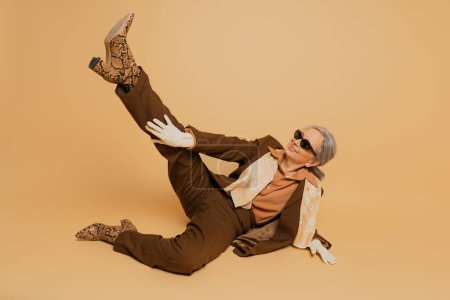 full length of happy senior woman in stylish sunglasses and suit posing with raised leg on beige background 
