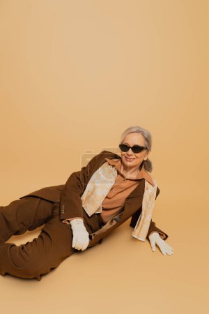 happy senior woman in stylish sunglasses and suit sitting and smiling on beige background 