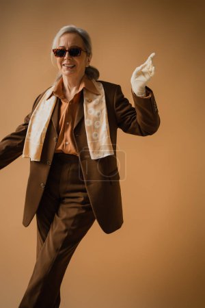Photo for Smiling senior woman in brown suit and white gloves gesturing on beige - Royalty Free Image