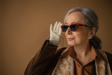 senior woman in brown suit and white gloves adjusting trendy sunglasses on beige 