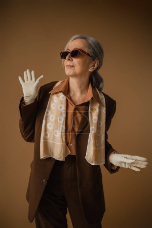 Photo for Senior woman in brown suit and white gloves gesturing and looking away on beige - Royalty Free Image