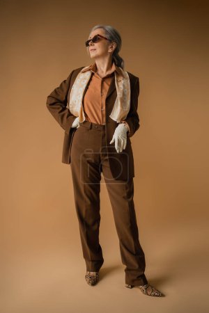Photo for Full length of senior woman in brown suit and white gloves posing with hand on hip on beige - Royalty Free Image