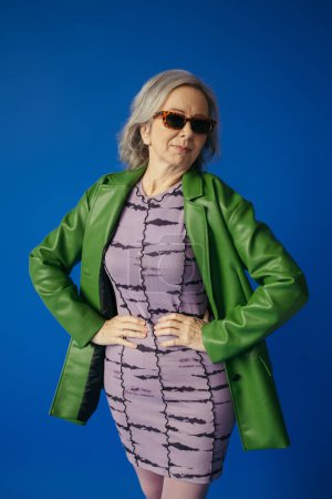 Seniorin in grüner Lederjacke und Kleid stehend mit Händen auf Hüften vereinzelt auf blauem Grund