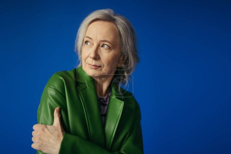 pensive grey haired woman in green leather jacket looking away isolated on blue