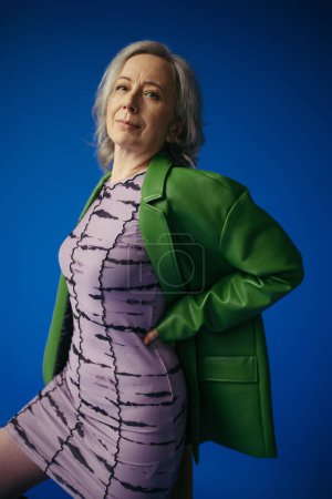 senior woman in elegant purple dress and green leather jacket posing with hand on waist isolated on blue