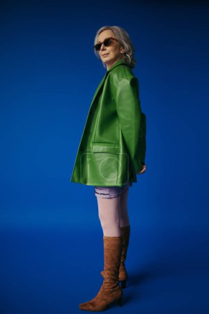 Téléchargez les photos : Longueur totale du mannequin senior en veste en cuir vert et lunettes de soleil élégantes debout sur fond bleu - en image libre de droit