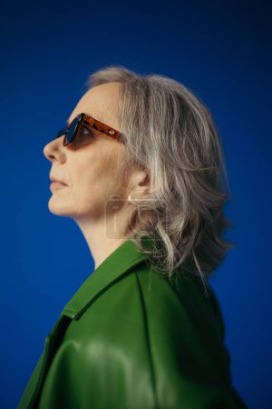 Photo for Profile of senior grey haired lady in trendy sunglasses and green leather jacket isolated on blue - Royalty Free Image