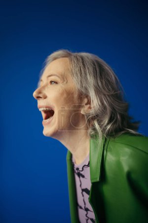 Foto de Astonished senior woman in green leather jacket looking away isolated on blue - Imagen libre de derechos