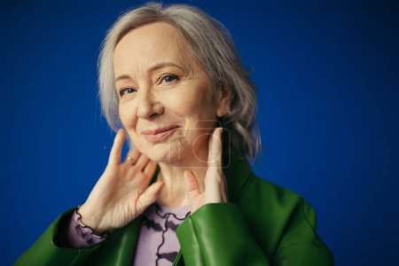 Porträt einer positiven und stilvollen Seniorin, die mit Händen nahe am Hals posiert und isoliert auf blauem Grund in die Kamera lächelt