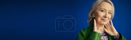 Photo for Portrait of smiling senior woman in green leather jacket posing with hands near neck isolated on blue, banner - Royalty Free Image