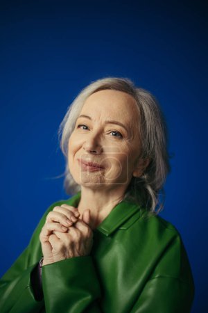 Porträt einer zufriedenen Seniorin in grüner Lederjacke, die isoliert auf blauem Grund in die Kamera blickt
