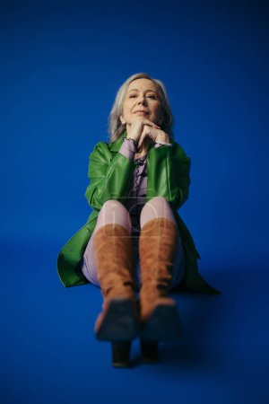 full length of trendy senior woman in green leather jacket and brown suede boots looking at camera on blue background