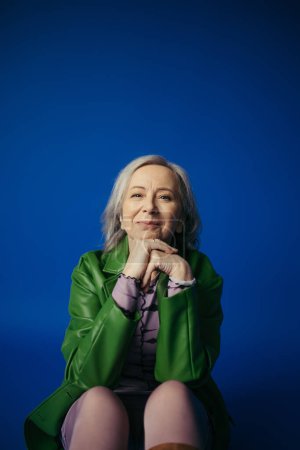 trendy senior woman in green leather jacket sitting with hands near face and smiling at camera on blue background