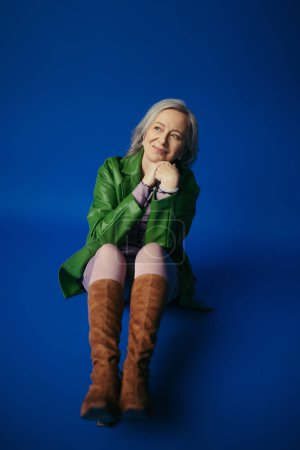 full length of dreamy and smiling senior woman in fashionable clothes sitting and looking away on blue background