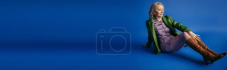 Foto de Full length of stylish grey haired woman in purple dress and green leather jacket sitting on blue background, banner - Imagen libre de derechos