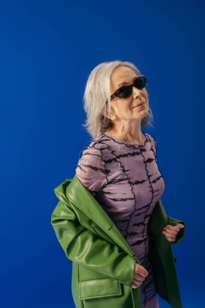 Seniorin mit Sonnenbrille und grüner Lederjacke über lila Kleid lächelt vereinzelt auf blau