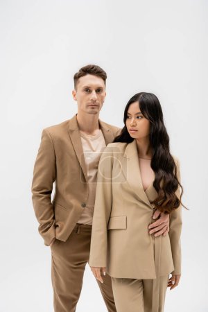 man in beige suit standing with hand in pocket and embracing asian woman isolated on grey