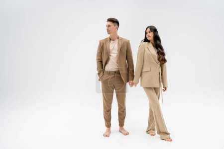 full length of barefoot interracial couple in beige pantsuits looking away on grey background