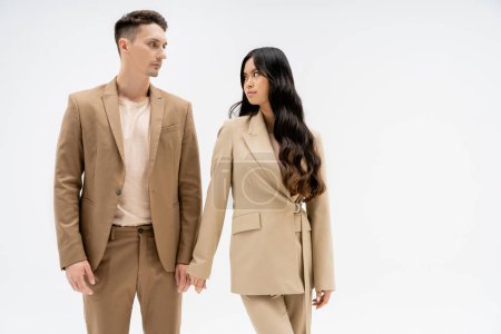 fashionable multiethnic couple in beige suits looking at each other isolated on grey