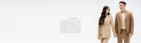 Téléchargez les photos : Élégant couple multiethnique en costumes beige debout et se regardant isolé sur gris, bannière - en image libre de droit