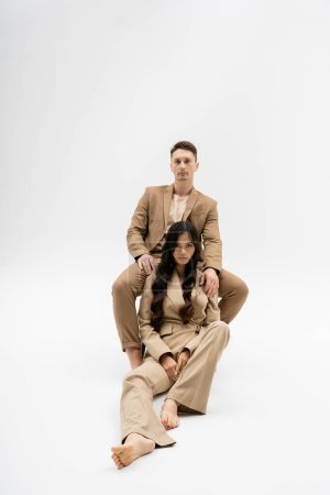 interracial couple in fashionable pantsuits looking at camera while posing on grey background