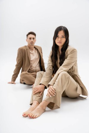 barefoot asian woman in beige pantsuit looking at camera near stylish man sitting on grey background