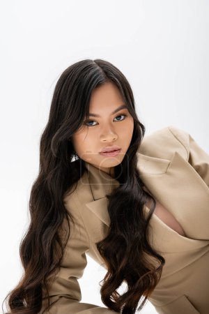 charming asian woman with long brunette hair looking at camera while posing in beige blazer on grey background
