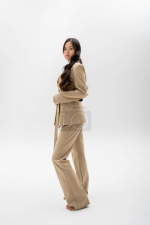 full length of barefoot asian woman in beige jacket and trousers standing on grey background