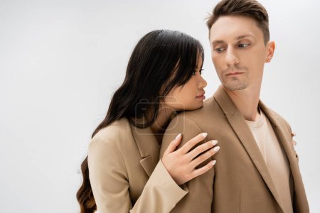 sensual asian woman with long brunette hair hugging man in beige jacket isolated on grey