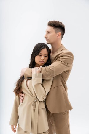 Téléchargez les photos : Trendy man in beige suit embracing asian woman with long brunette hair on grey background - en image libre de droit