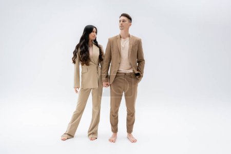 full length of barefoot multiethnic couple in trendy suits holding hands while standing on grey background