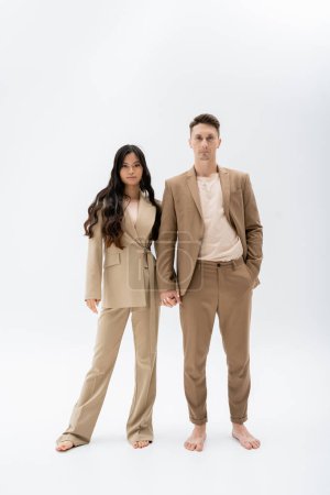 longitud completa de la pareja multiétnica descalza en trajes de pantalón beige tomados de la mano sobre fondo gris