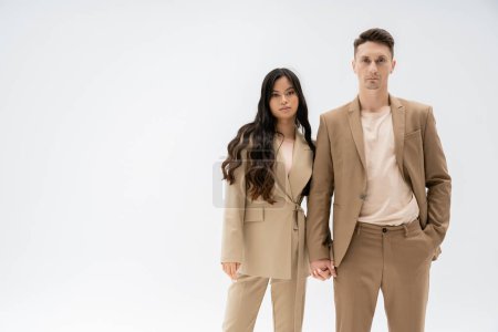 pareja interracial en traje de moda tomados de la mano y mirando a la cámara aislada en gris