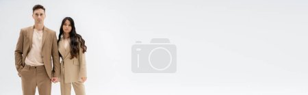 Téléchargez les photos : Couple multiethnique branché en costumes beige regardant la caméra et tenant les mains isolées sur gris, bannière - en image libre de droit