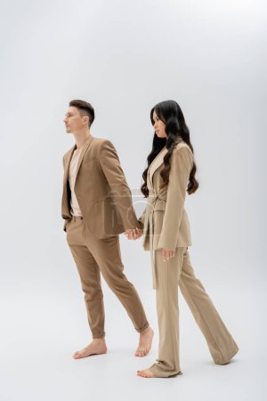 full length of barefoot multiethnic couple in beige pantsuits holding hands while walking on grey background