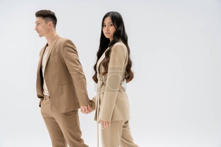 young asian woman with long hair looking at camera while holding hands with man in beige suit isolated on grey