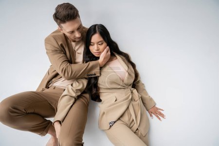 high angle view of interracial romantic couple in beige suits touching each other while posing on grey background