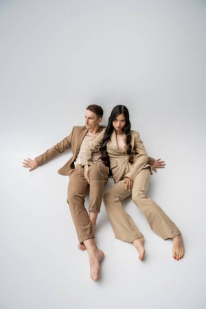 longitud completa de la pareja multiétnica descalza en traje de moda sentado sobre fondo gris