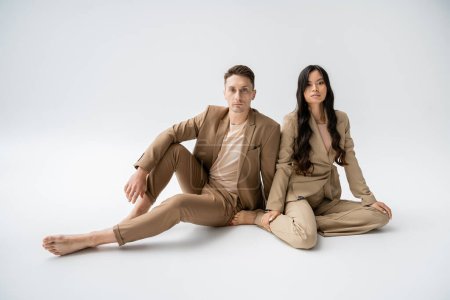 full length of barefoot interracial couple in stylish attire sitting and looking at camera on grey background
