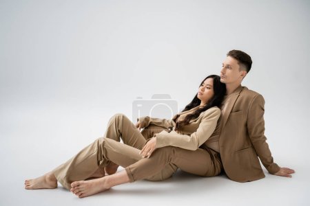 full length of barefoot interracial couple in beige pantsuits sitting on grey background