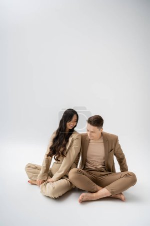 happy interracial couple in stylish pantsuits sitting with crossed legs on grey background