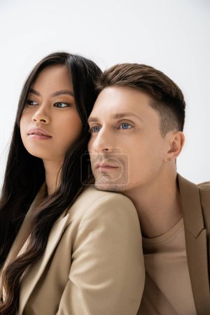 stylish man leaning on shoulder of brunette asian woman and looking away isolated on grey