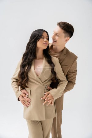 Téléchargez les photos : Smiling man hugging brunette asian woman in trendy casual attire isolated on grey - en image libre de droit